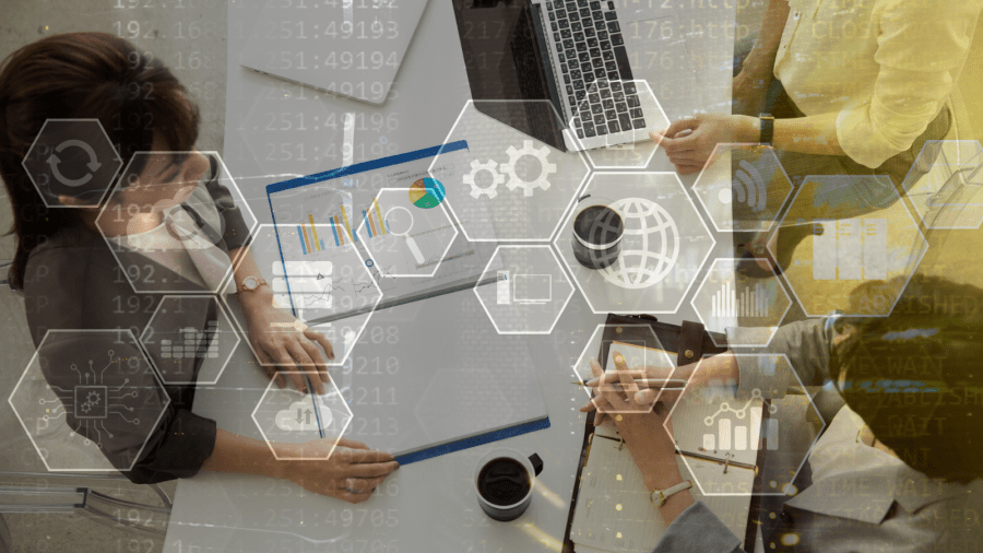 Three professionals collaborate at a desk with laptops, charts, analyzing data and discussing strategy. Transparent hexagonal icons representing technology, networking, and analytics overlay the image.