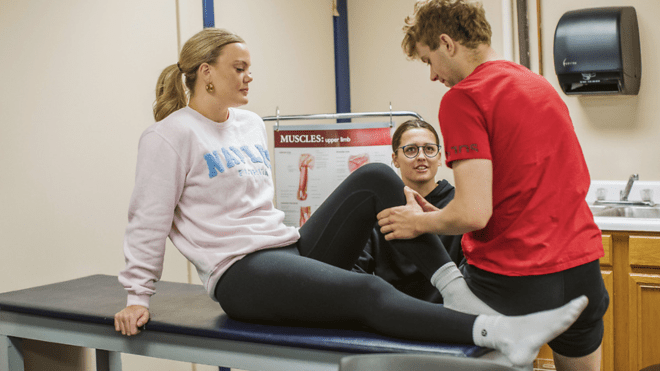 Student performing a knee exam on another student in a physical therapy classroom with a teacher observing 