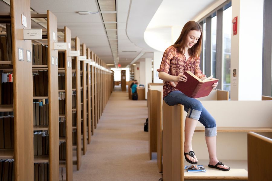 A student sitting on a desk reading a book near a large amount of other books on shelves
