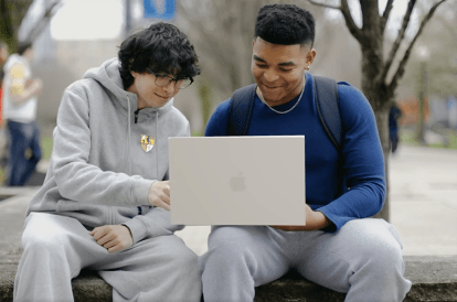 2 students sitting outside on a college campus looking at a laptop together