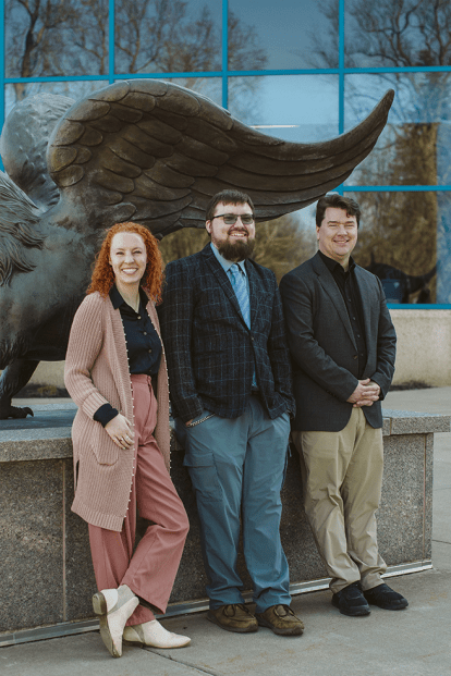3 college staff members posing on a street in front of a statue of a Griffin